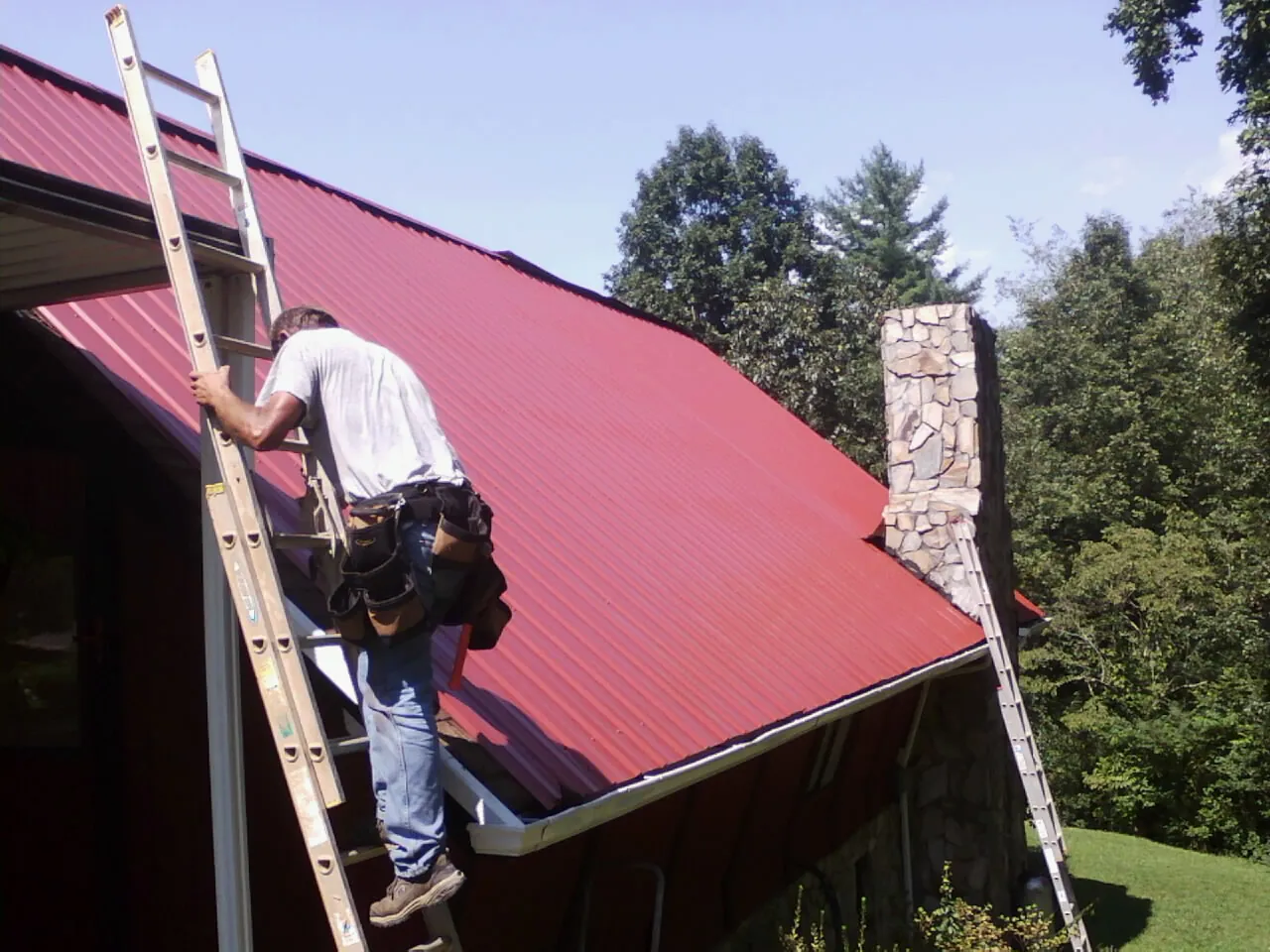 Roofer on a ladder beside a red metal roof and stone chimney — Soffit & Fascia Repair by Roofing Brothers in Fox River Grove